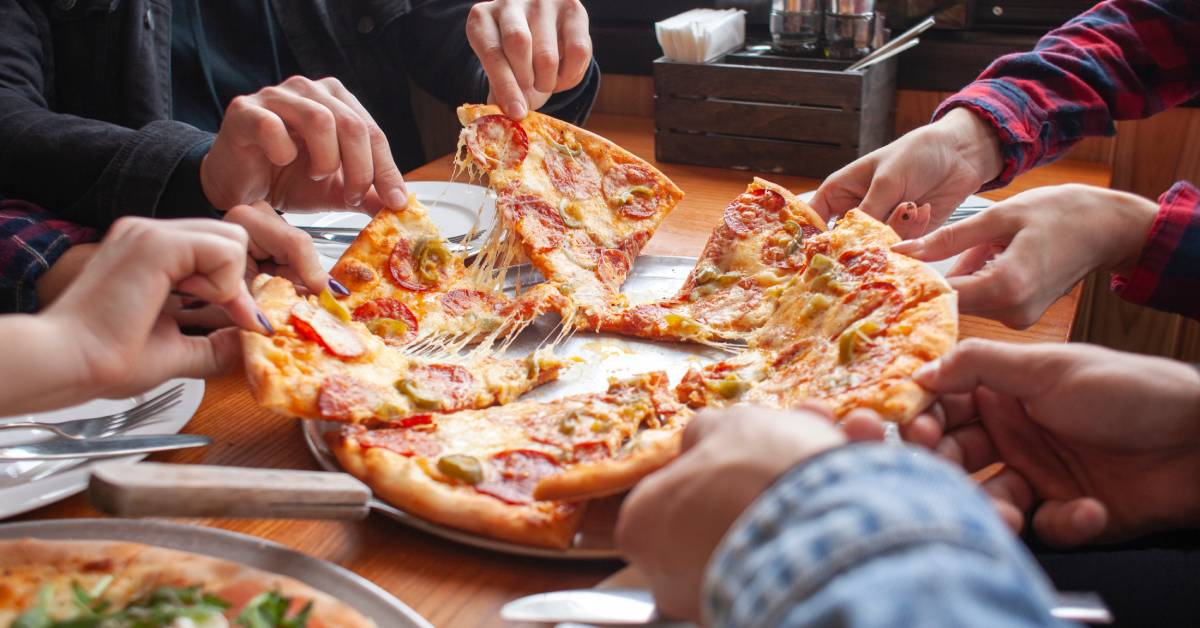 A group of people reaching for slices of pizza at a communal table—the pizza is topped with pepperoni and jalapenos.