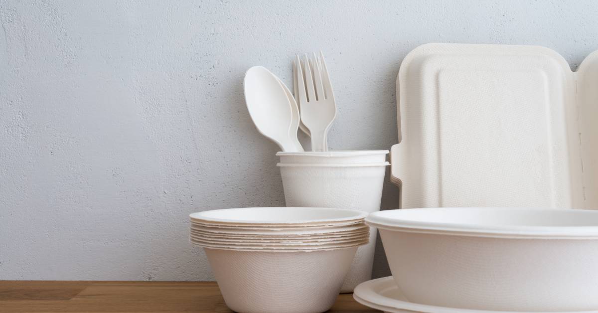 Sets of natural, eco-friendly disposable utensils and food containers made from bamboo on a wooden table in front of a white wall.