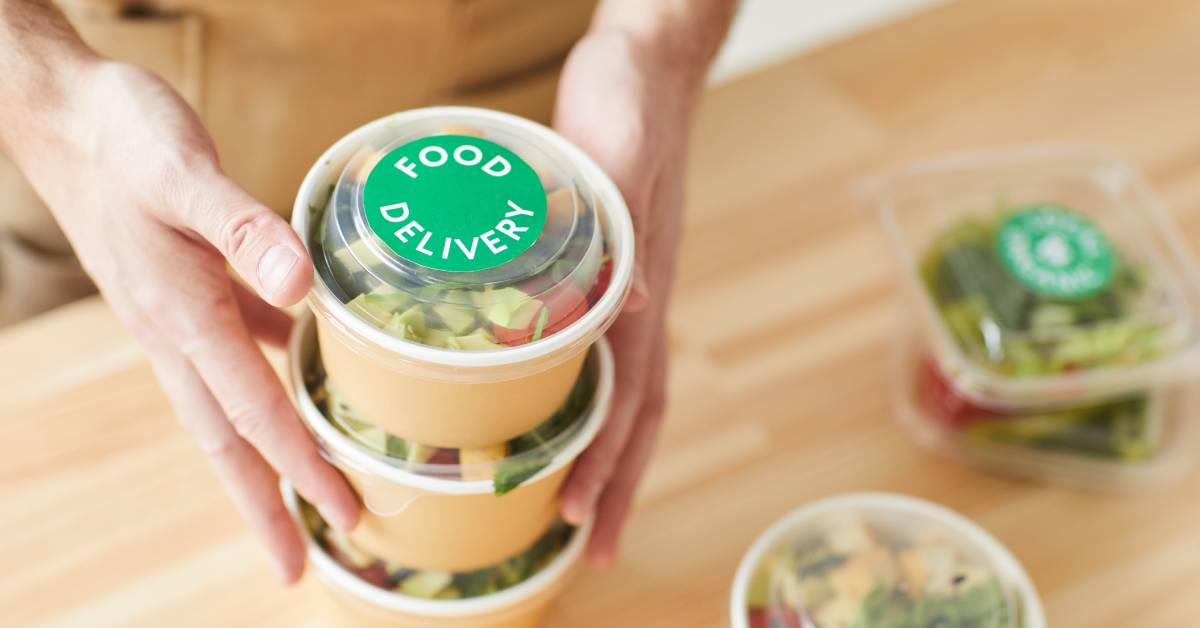 High-angle close-up on a stack of food delivery containers full of salad with more on the table below, made of cardboard.