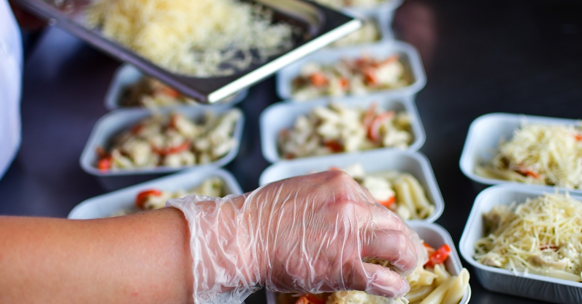 Dos and Don’ts When Sourcing Catering Boxes A close-up view shows a person wearing a food-safety glove placing food into a cardboard food container.