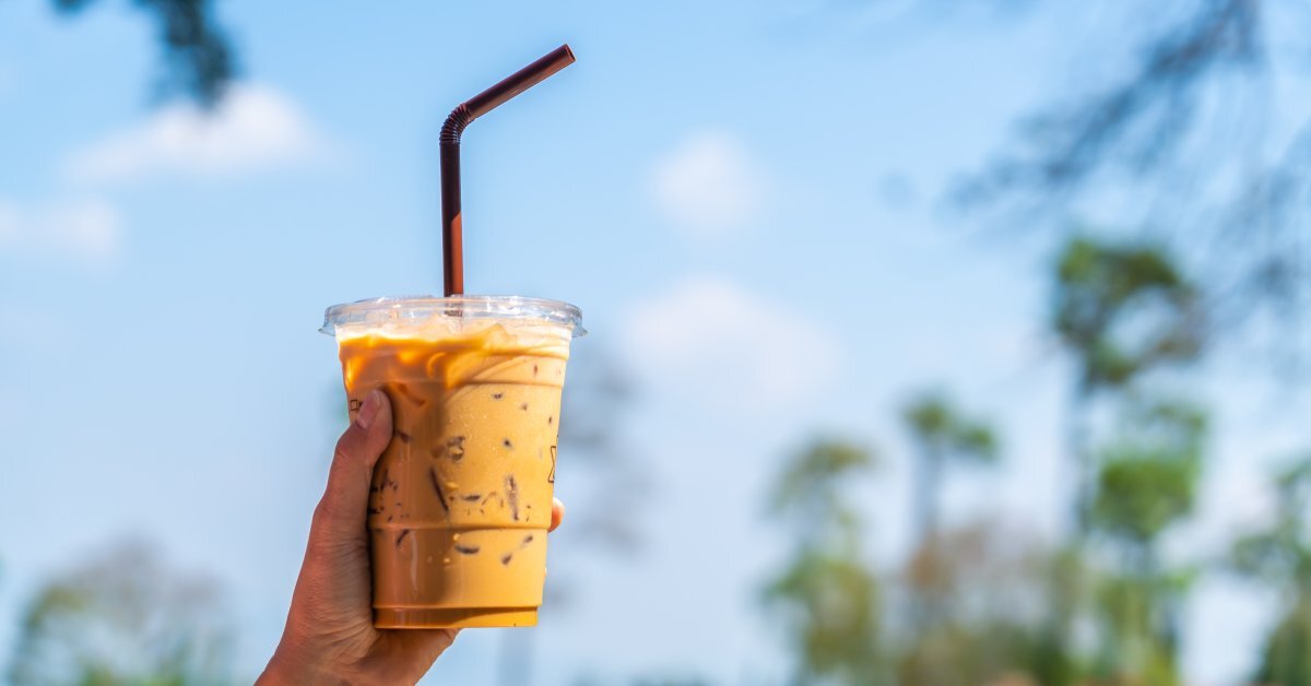 A hand holds an iced coffee cup with a straw against a blue sky, with blurred green foliage in the background.