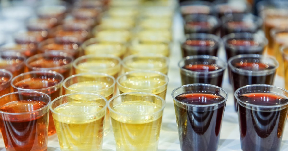 Rows of clear PET cups filled with colorful beverages, including amber, yellow, and dark red drinks, are on a white table.