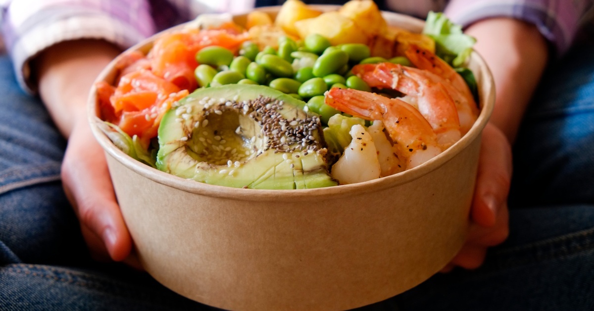 A person holds a paper take out container filled with shrimp, avocado, edamame, and salmon, showcasing a colorful, healthy meal.
