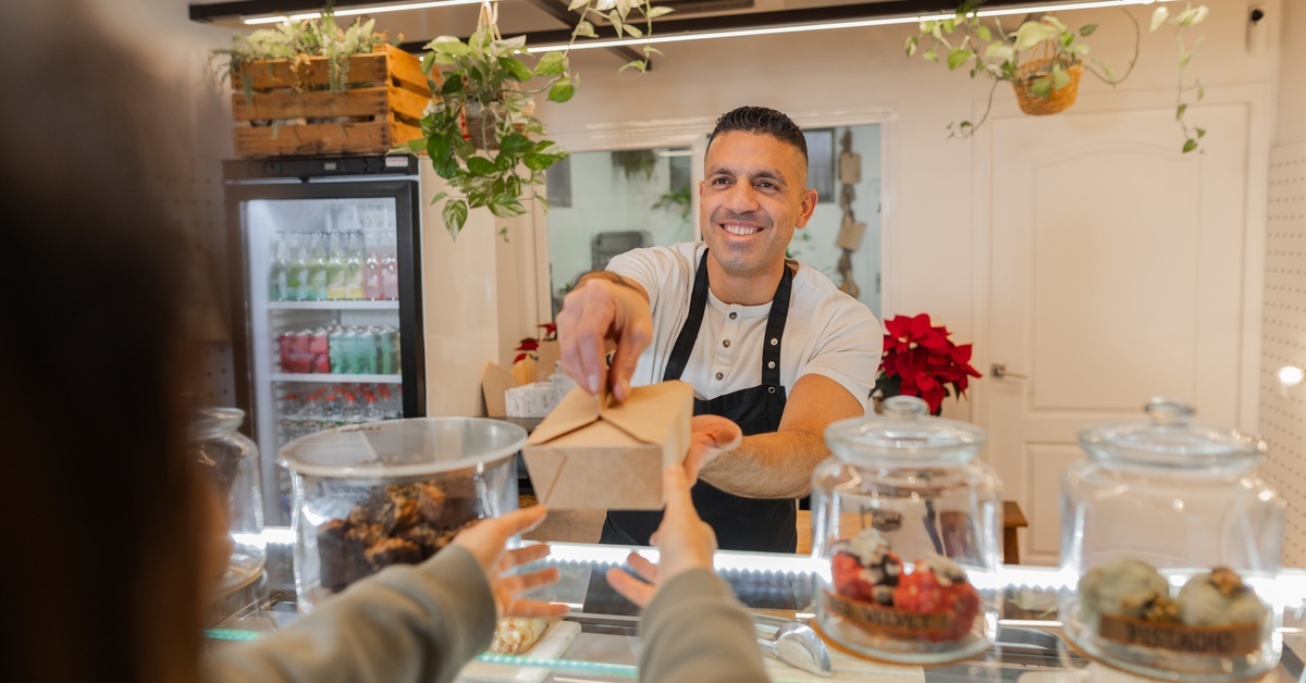 A smiling baker in a black apron hands a takeout box to a customer across the counter, containing bakery displays. A smiling baker in a black apron hands a takeout box to a customer across the counter, containing bakery displays.