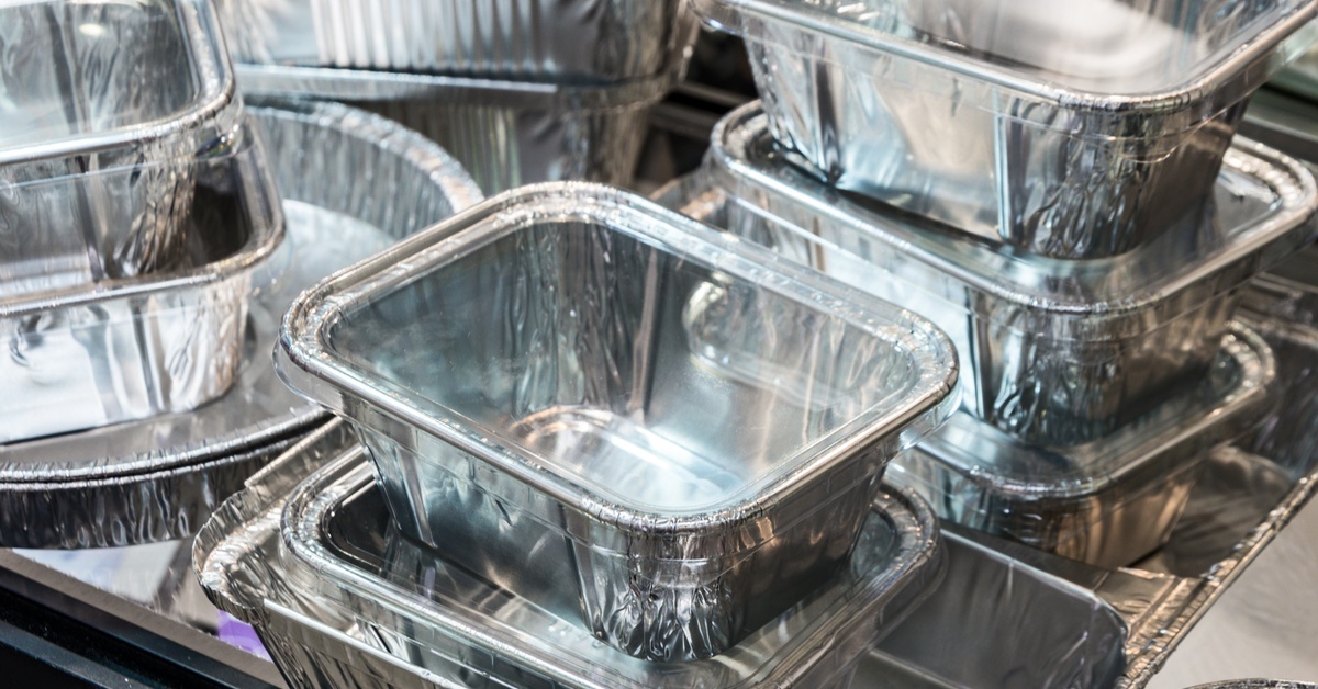 Small aluminum food containers with clear lids rest stacked together on a steel surface in a commercial kitchen.