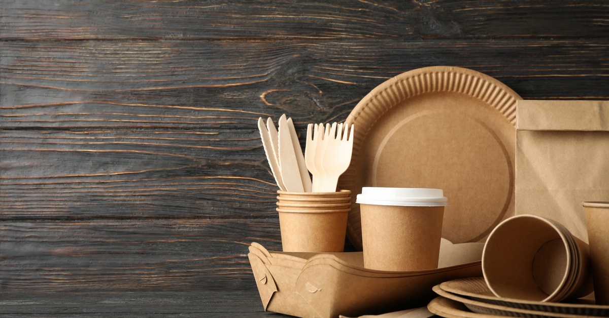 A wooden table full of cardboard tableware and one brown paper bag. The wall is also made of dark brown wood.