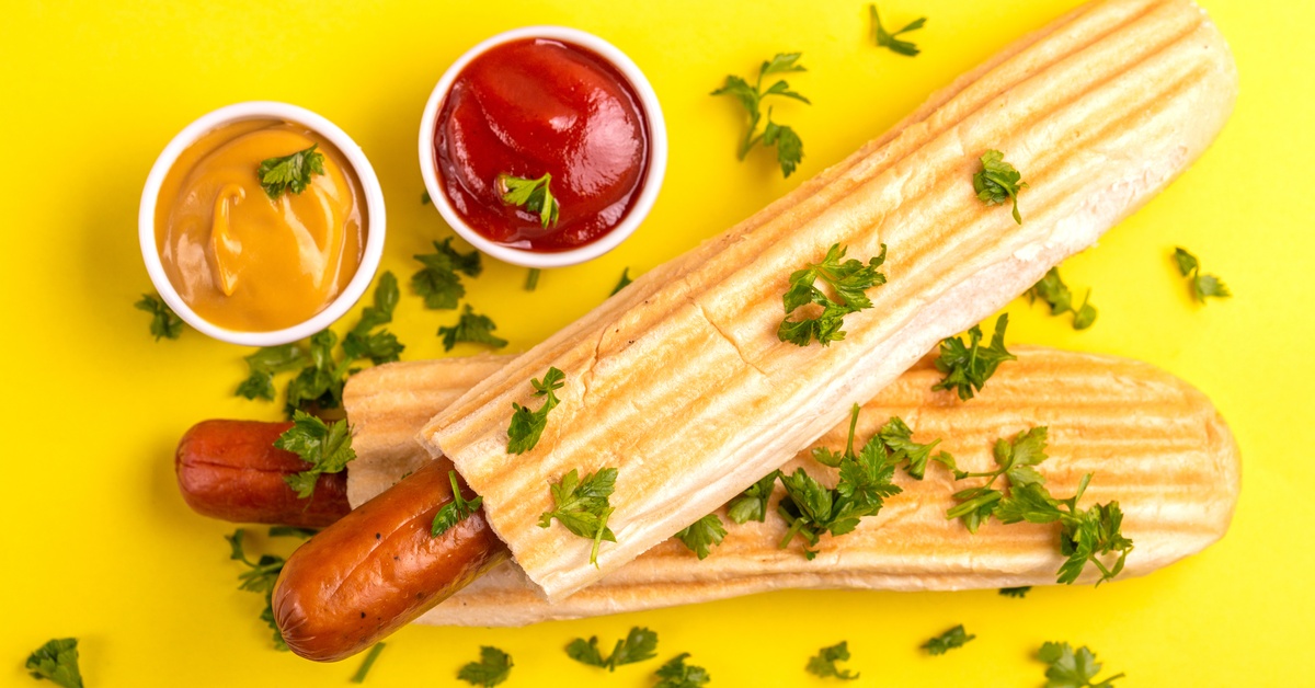 Grilled French hot dogs with mustard and ketchup in white portion cups on a bright yellow background with fresh herbs.