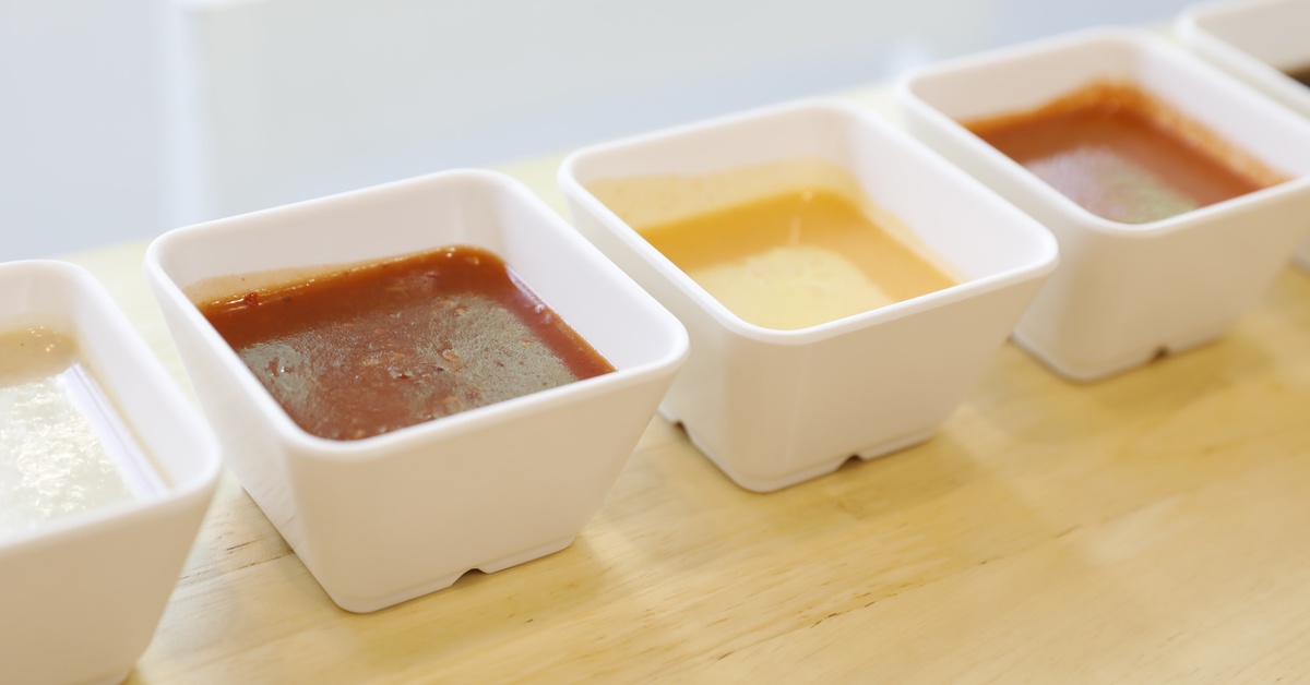 Multiple white square portion cups, each filled with a different sauce or condiment, line up on a wooden serving board.