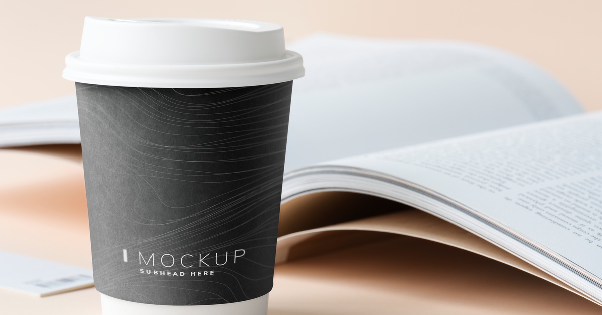 A black custom cup with a white lid displays the text "MOCKUP SUBHEAD HERE" beside an open book on a neutral background.