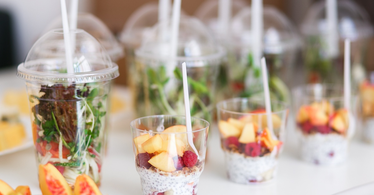 Clear custom cups filled with colorful fruit, chia pudding, and fresh greens with white forks on a display counter.