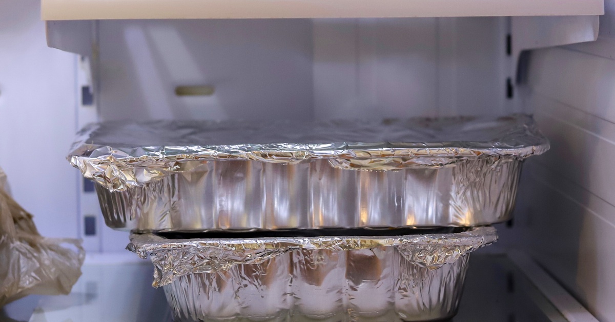Two aluminum pans full of food with aluminum foil on top sit in a refrigerator stacked together.