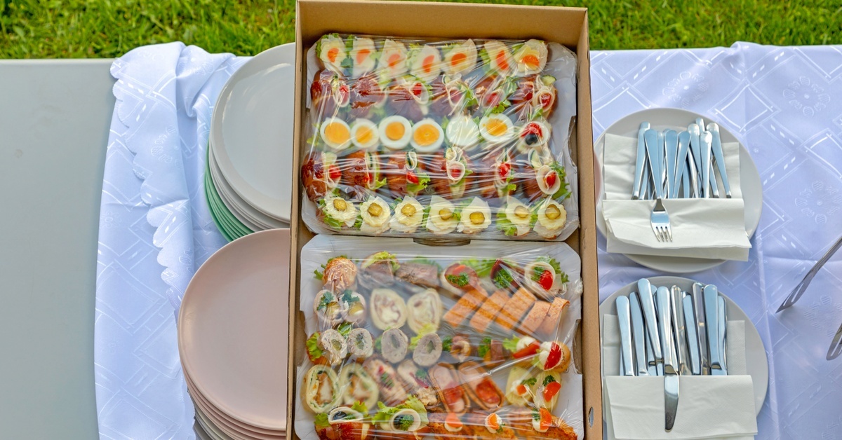 A paper catering box filled with an assortment of sushi and wraps on an outdoor table set with plates and silverware.
