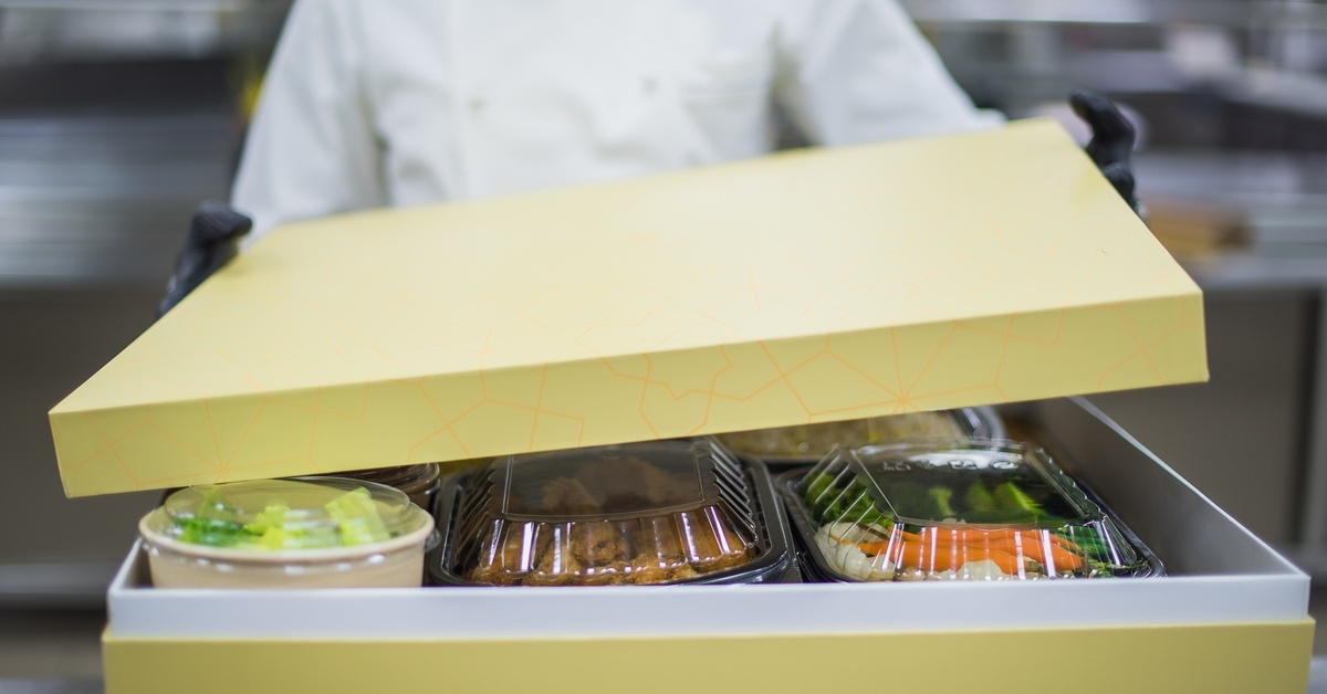 A chef opens a catering box with plastic containers filled with prepared meals in a professional commercial kitchen.