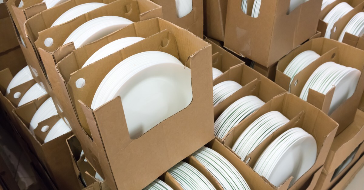 Stacks of white and round-shaped porcelain plates in boxes are on the floor of a restaurant storage facility.
