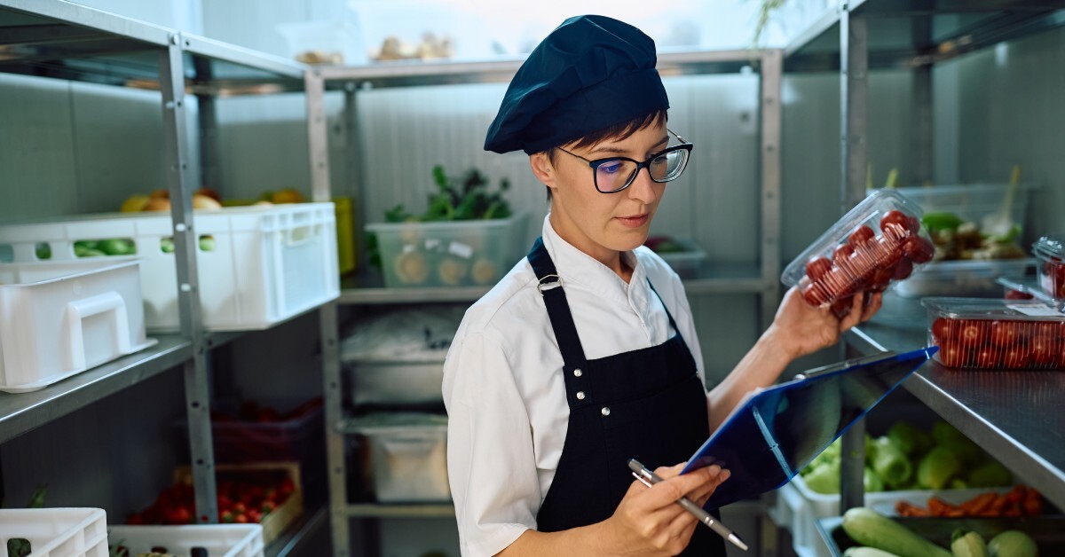 A female chef wearing a black hat, a black apron, and a white shirt uses a clipboard to check restaurant food supplies.