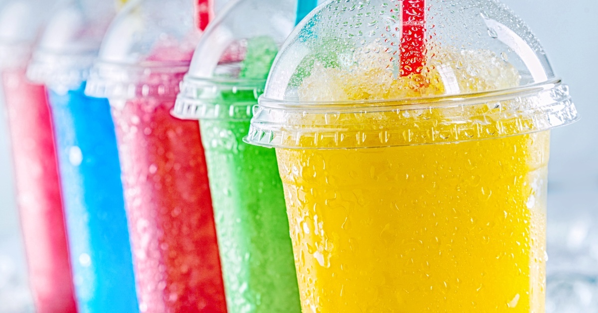 Colorful frozen drinks in clear cups with dome lids and straws sit on ice, showing yellow, green, red, and blue slushes.