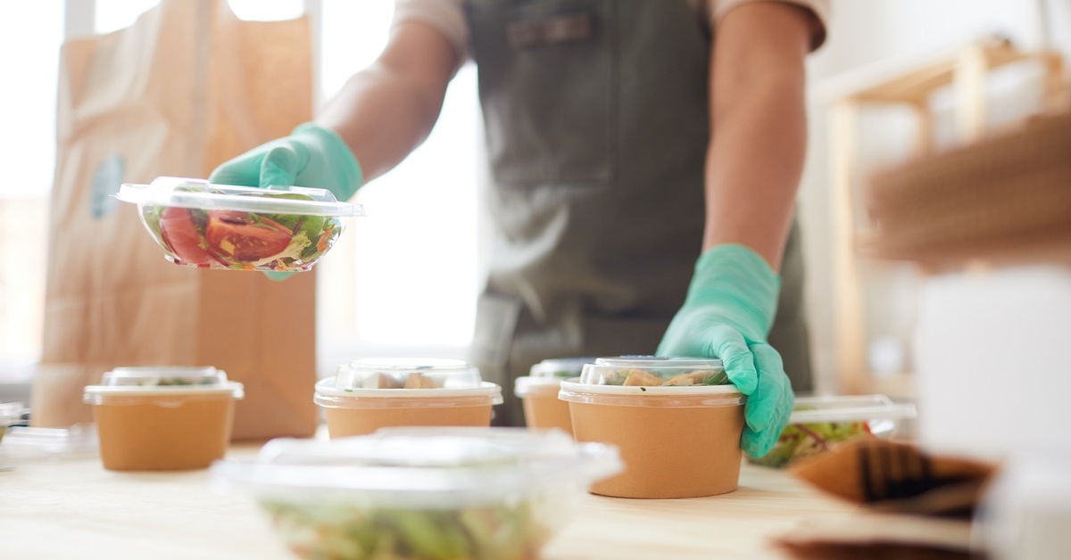 Foodservice worker wearing gloves packs fresh salads into plastic takeout containers on a prep table in a commercial kitchen.