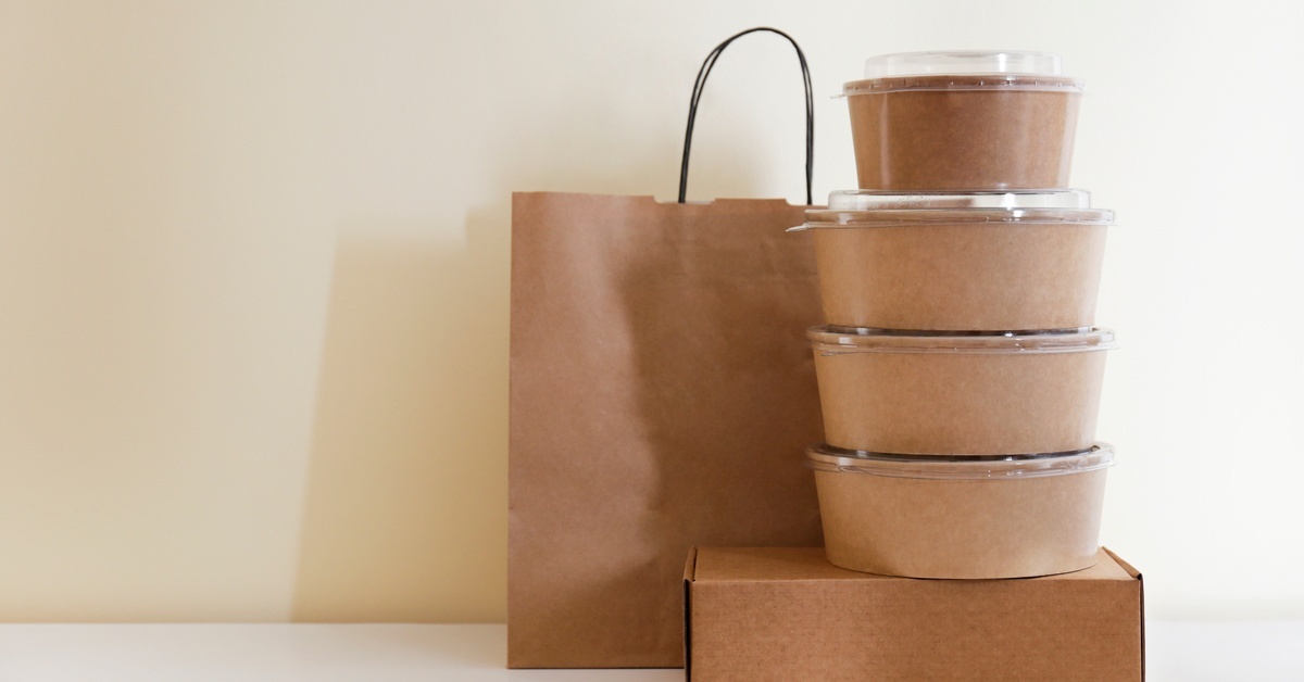 Stacked kraft takeout bowls with clear lids sit beside a paper carry bag and box on a clean, neutral surface.