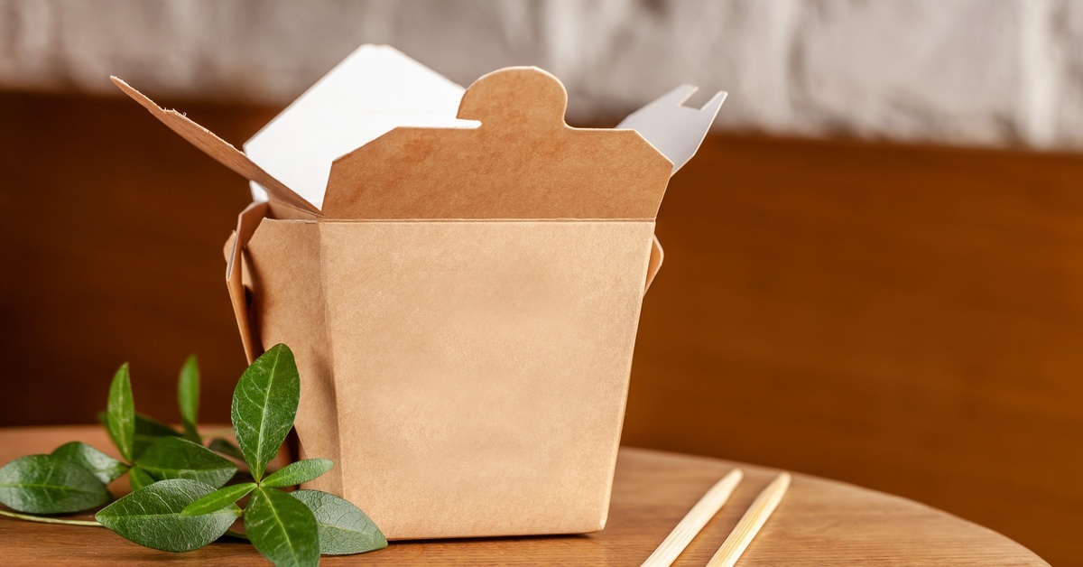 A Kraft paper take-out box with a folded top sits on a wooden table beside chopsticks and green leaves.