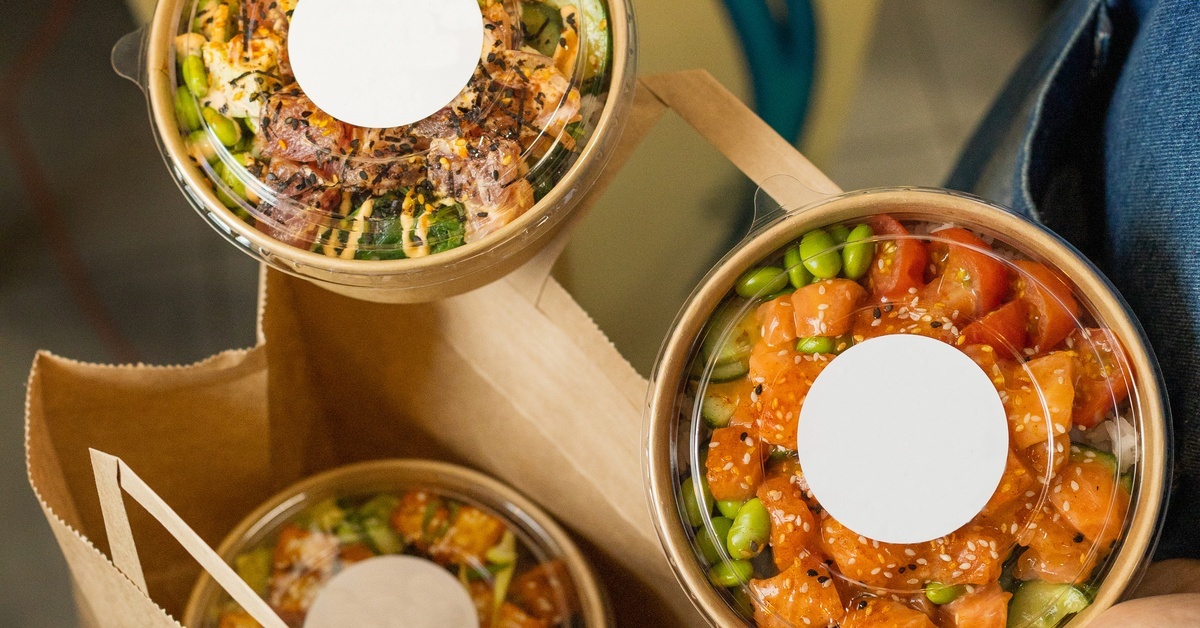 A person holds clear take-out bowls with poke and vegetables beside a paper bag filled with additional take-out containers.