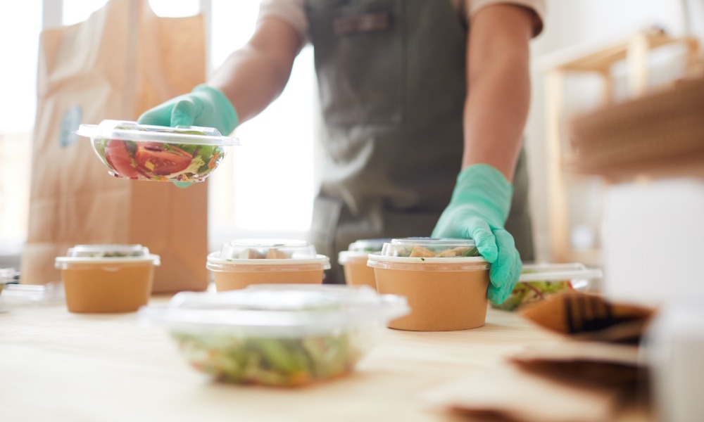 A foodservice worker wearing gloves packing fresh salads into sealed takeout containers on a prep table for leak-resistant takeout.