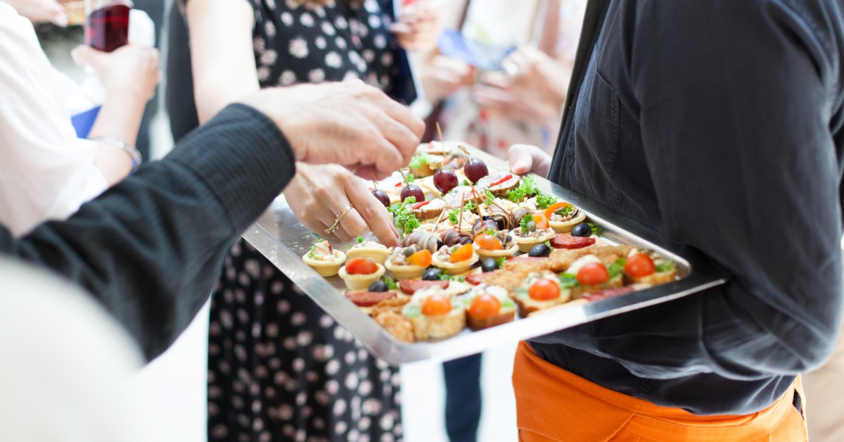 Guests select assorted bite-sized appetizers from a catering tray at a social event with drinks in hand.