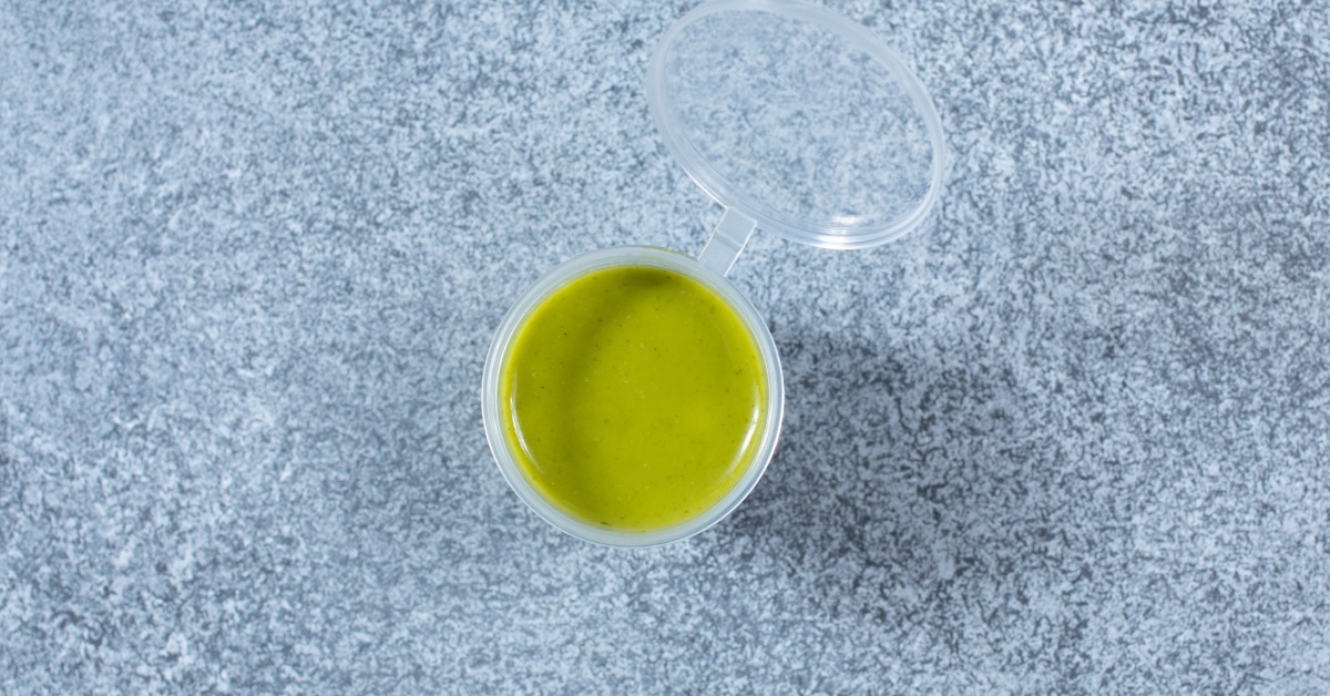 A clear plastic sauce cup with a hinged lid holds green dressing or sauce on a textured gray surface.