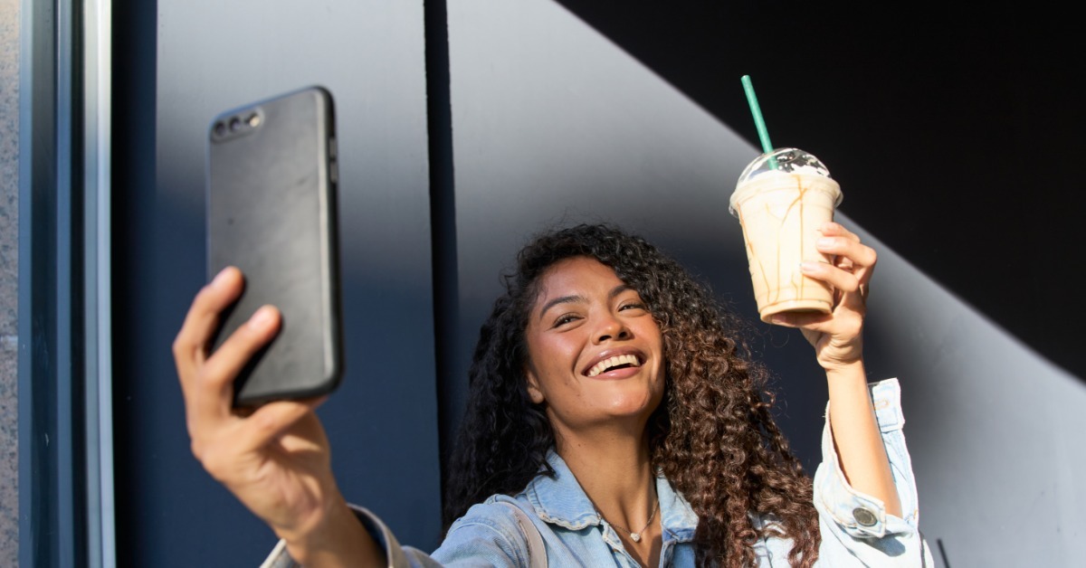A woman takes a selfie while holding a caramel iced coffee in a clear plastic cup with a dome lid and straw.