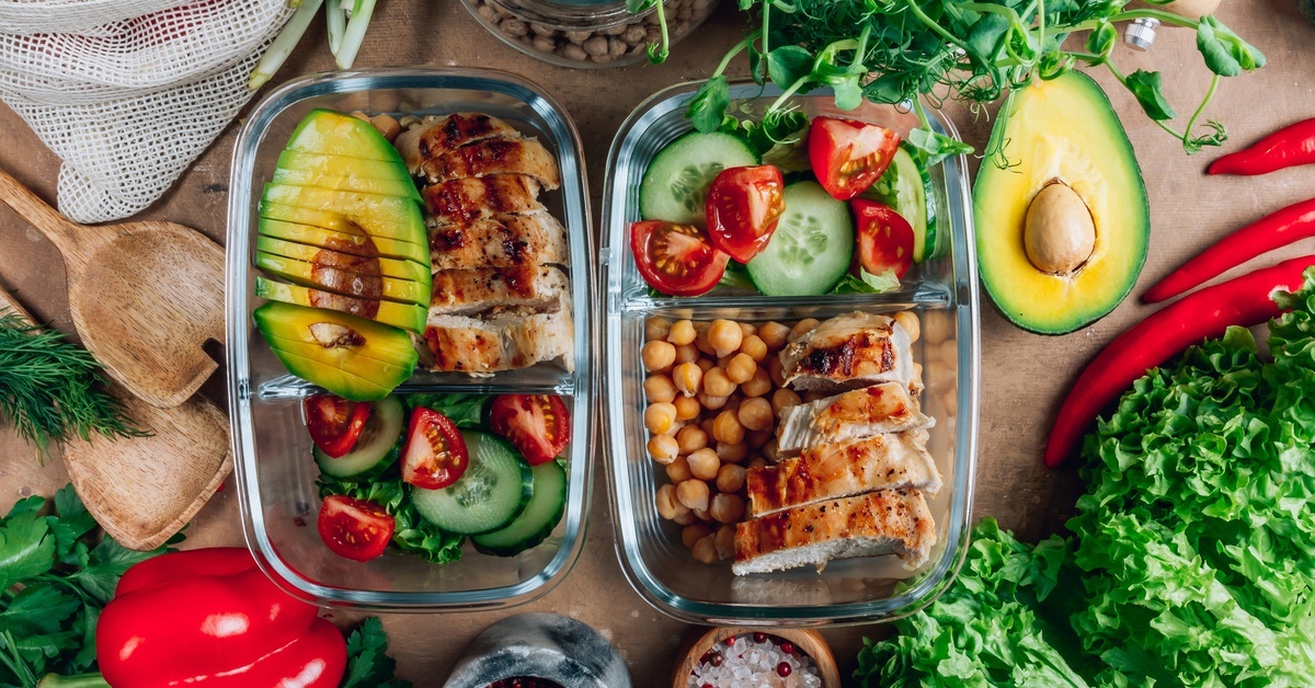 Glass meal prep containers with grilled chicken, avocado, chickpeas, and fresh vegetables arranged on a wooden surface.