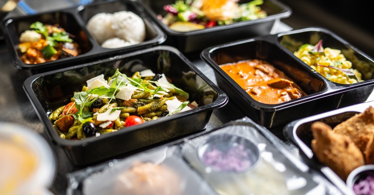 Black meal prep containers filled with assorted prepared meals, including salad, curry, and sides, arranged on a kitchen surface.