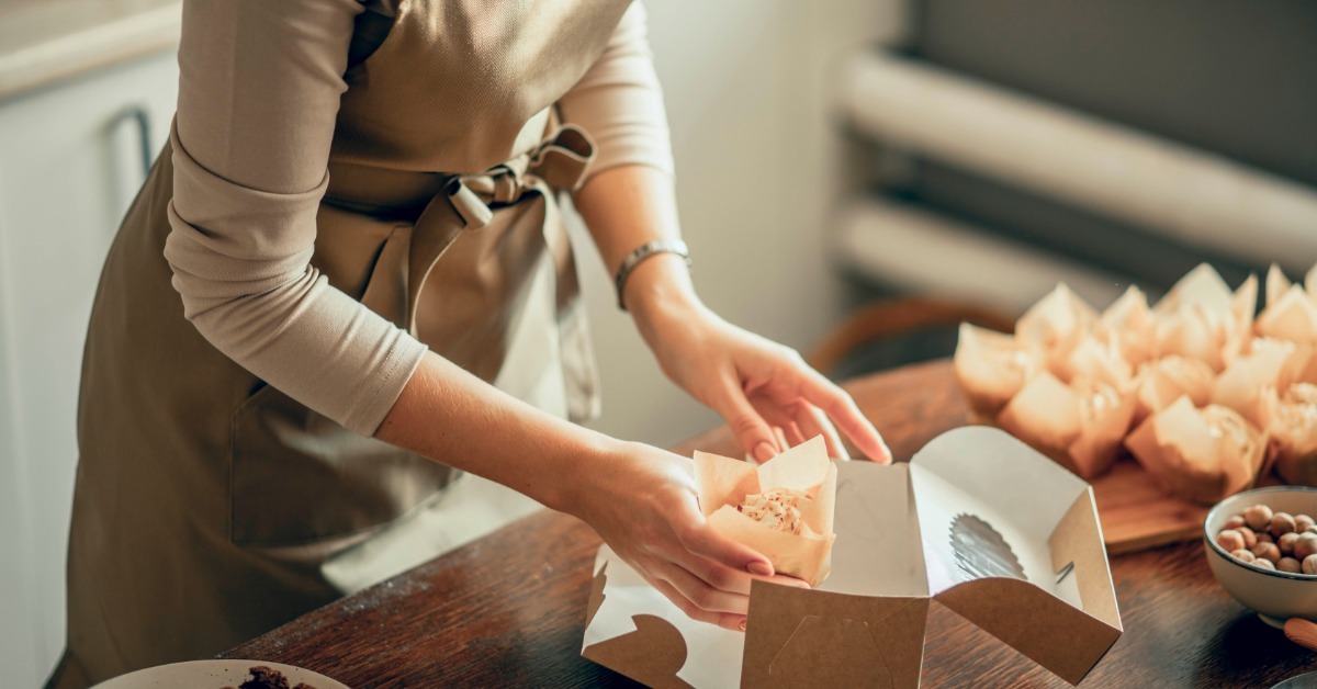 A bakery owner places wrapped cupcakes into a foldable box, showing efficient packaging and secure handling for delivery orders.