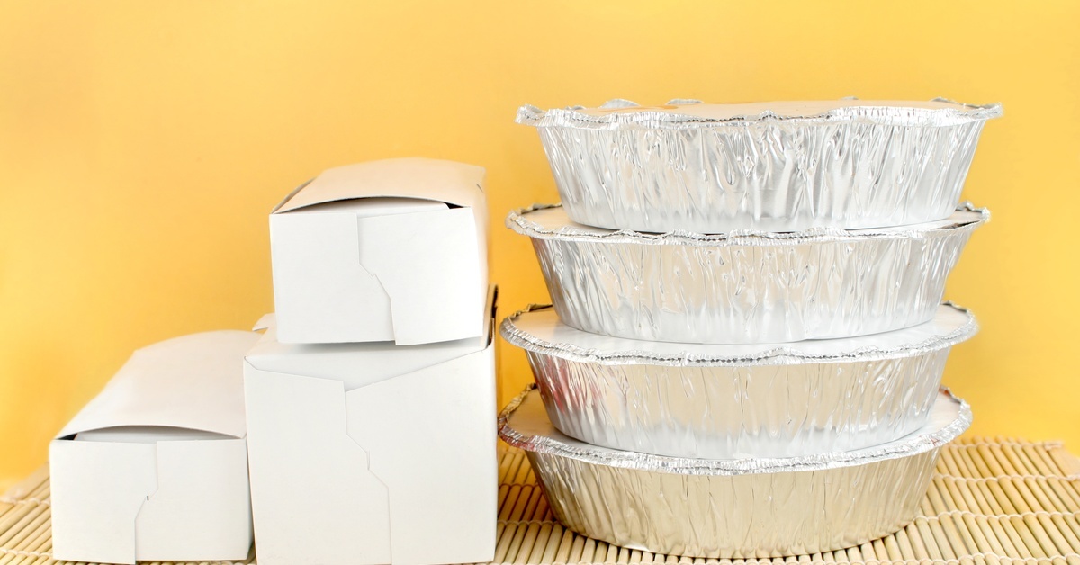 Four round aluminum foil containers stacked next to white cardboard takeout boxes on a bamboo mat with a yellow background.