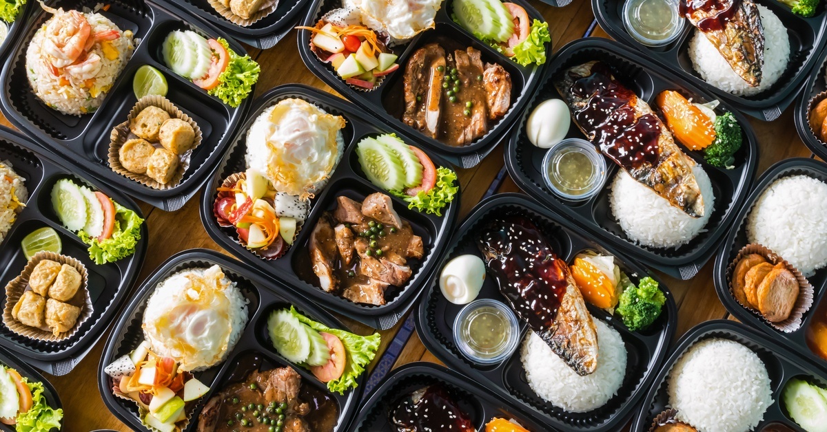 A variety of catered box meals with rice, grilled fish, eggs, vegetables, and sauces arranged in black compartment trays.