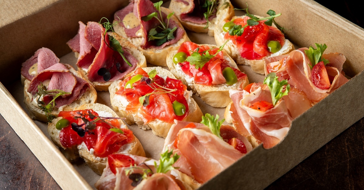 Assorted gourmet crostini with cured meats, smoked salmon, herbs, and olives arranged neatly in a kraft catering box.