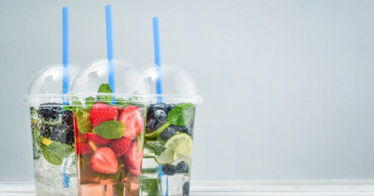 Three fruit-infused beverages in clear plastic dome-lid cups with blue straws and fresh berries displayed on a white wood surface.