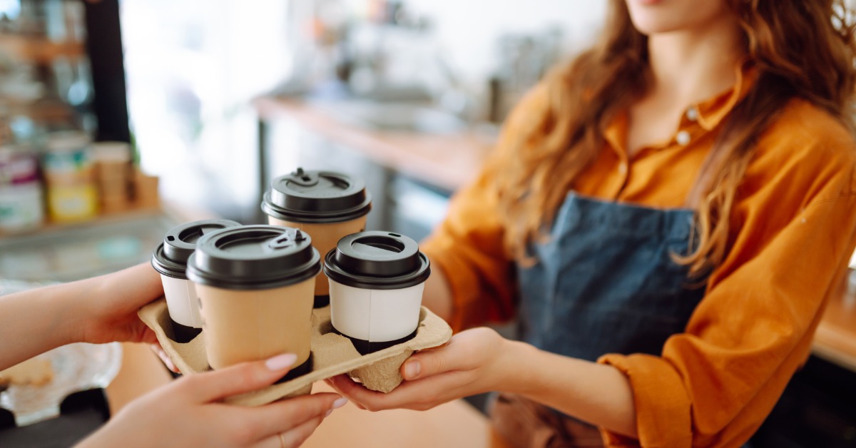 A barista hands a customer a cardboard carrier holding four paper to-go cups with black sipper lids inside a caf&eacute;.