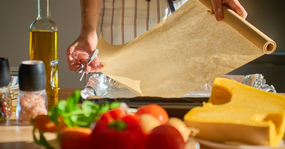 A cook in an apron unrolling parchment paper over a baking pan, with fresh vegetables, cheese, and olive oil nearby on the counter.