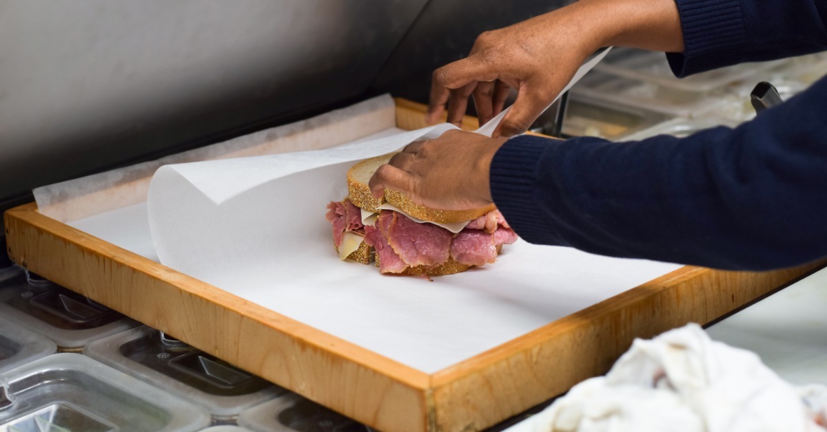 A deli worker wrapping a thick corned beef sandwich in wax paper on a wooden cutting board in a commercial kitchen.