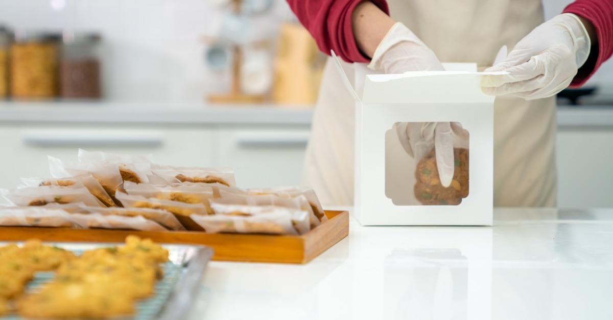 A baker wearing gloves places a cookie into a white window bakery box on a kitchen counter with packaged cookies nearby.