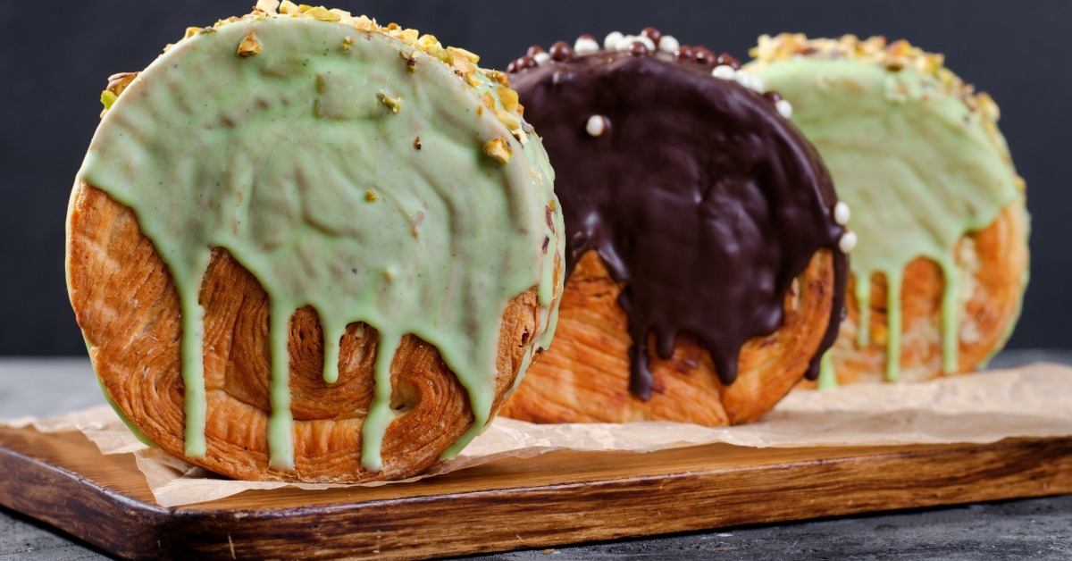 Round croissants with dripping pistachio and chocolate glaze topped with crushed nuts on parchment over a rustic wooden board.