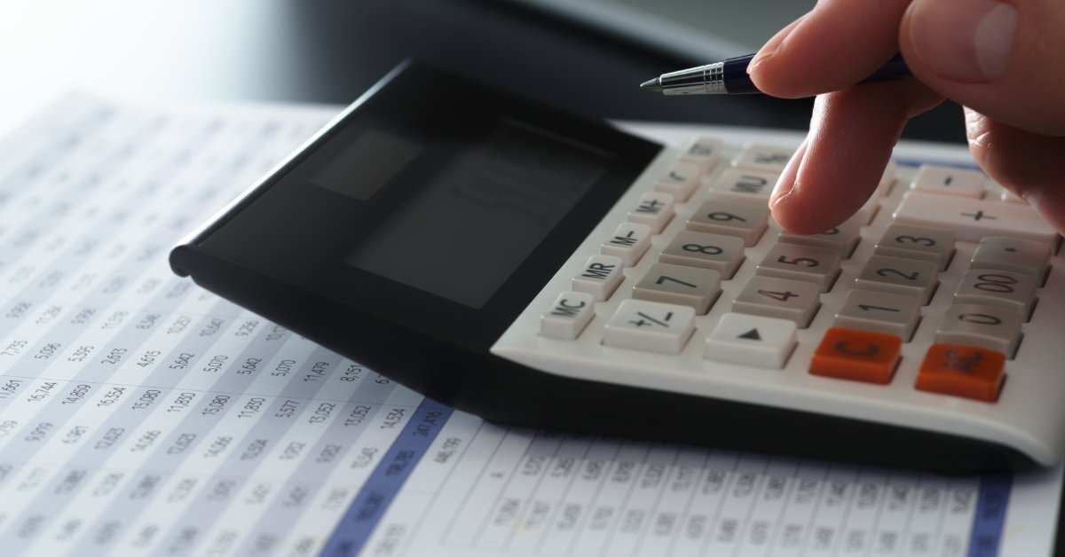 A close-up of a hand holding a pen while pressing keys on a desktop calculator over a financial spreadsheet.