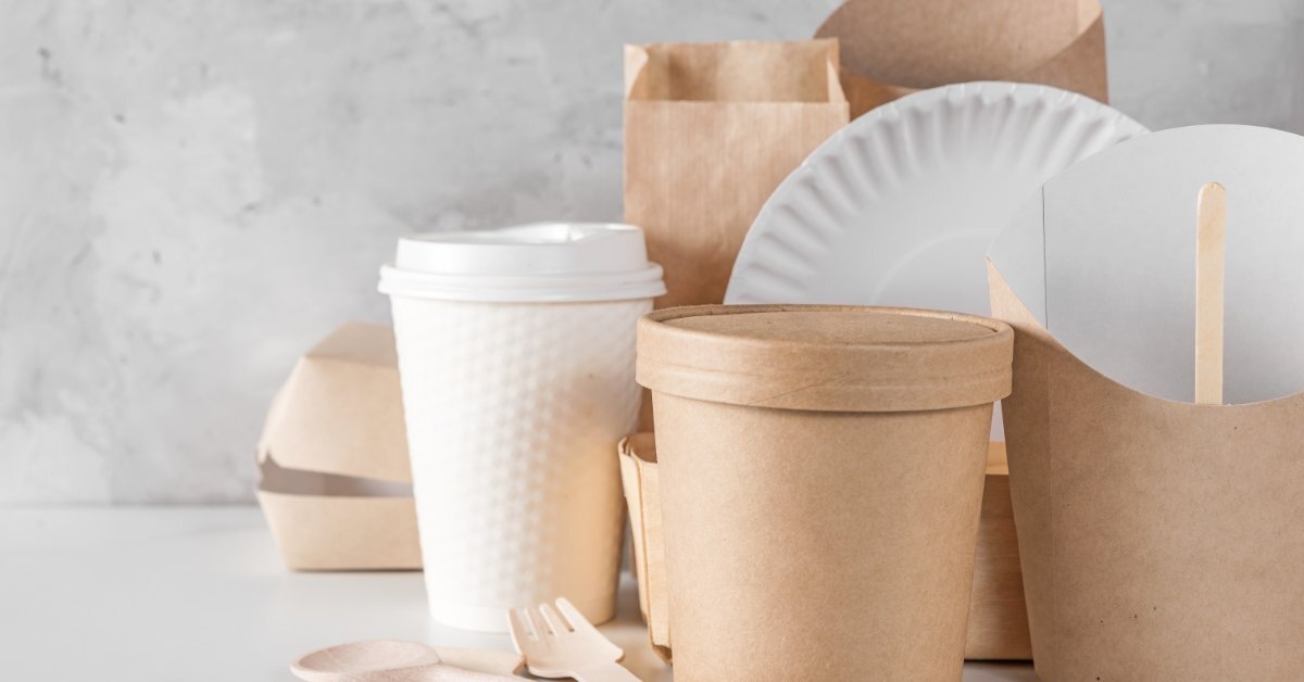 A collection of kraft paper cups, takeout containers, paper plates, and wooden cutlery arranged on a white tabletop.