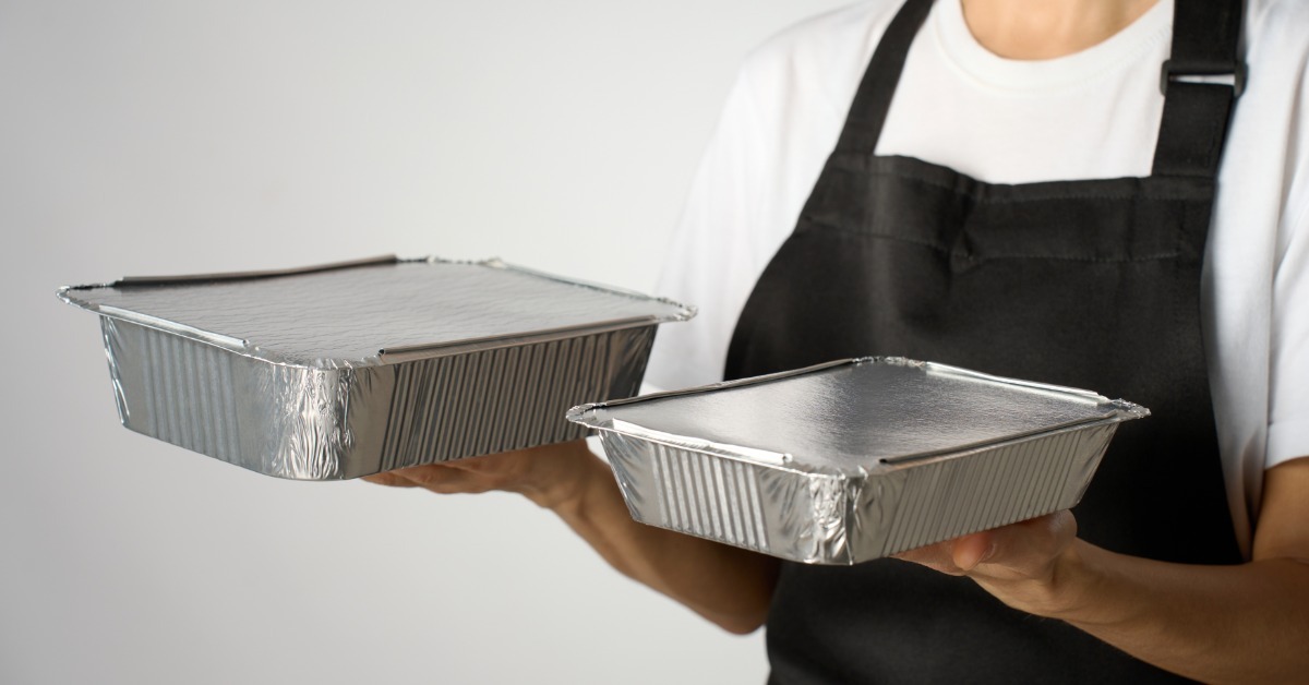 A food service worker in a black apron holds two aluminum foil catering containers against a light gray background.