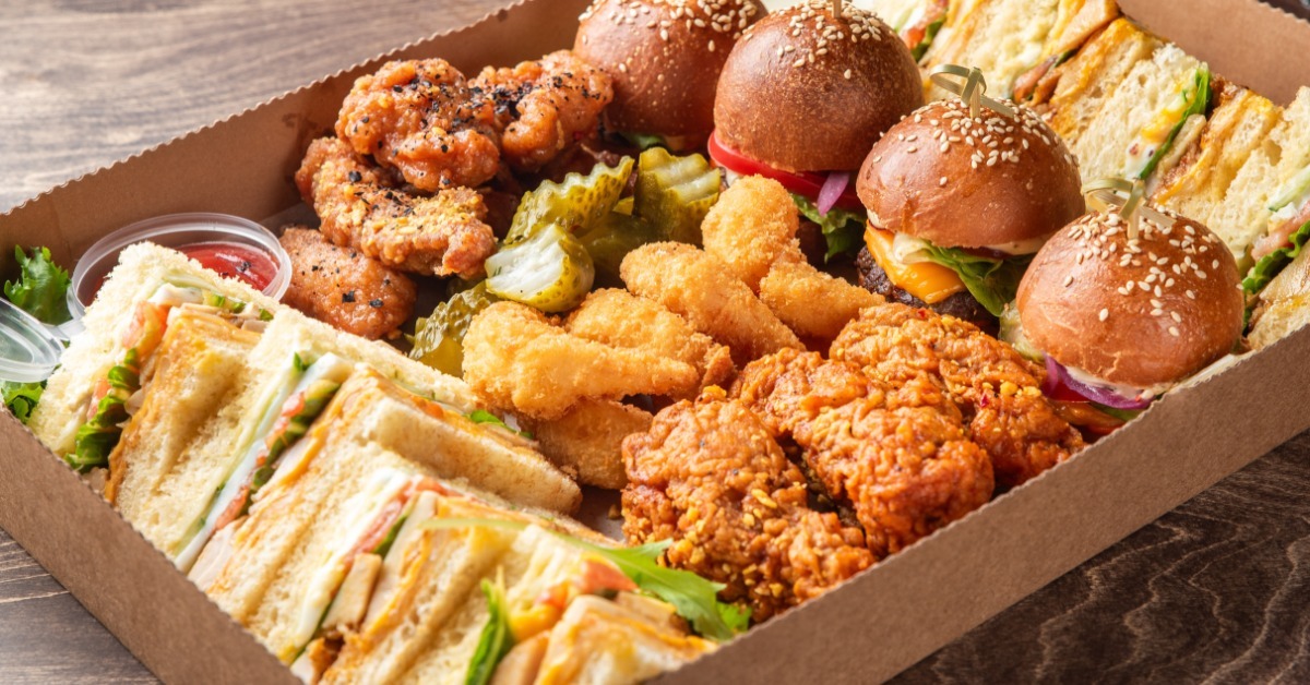 A kraft cardboard catering box filled with sliders, fried chicken, sandwiches, and sides on a wooden surface.