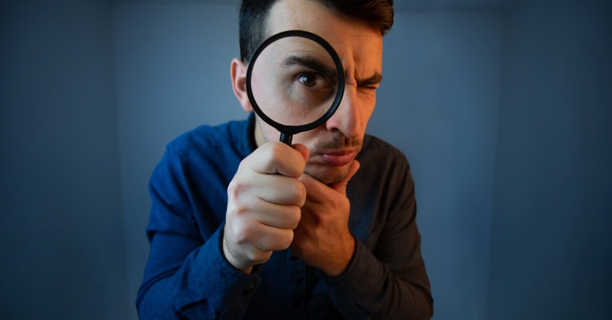 A man in a blue shirt holds a magnifying glass up to his eye with a serious expression against a blue background. A man in a blue shirt holds a magnifying glass up to his eye with a serious expression against a blue background.