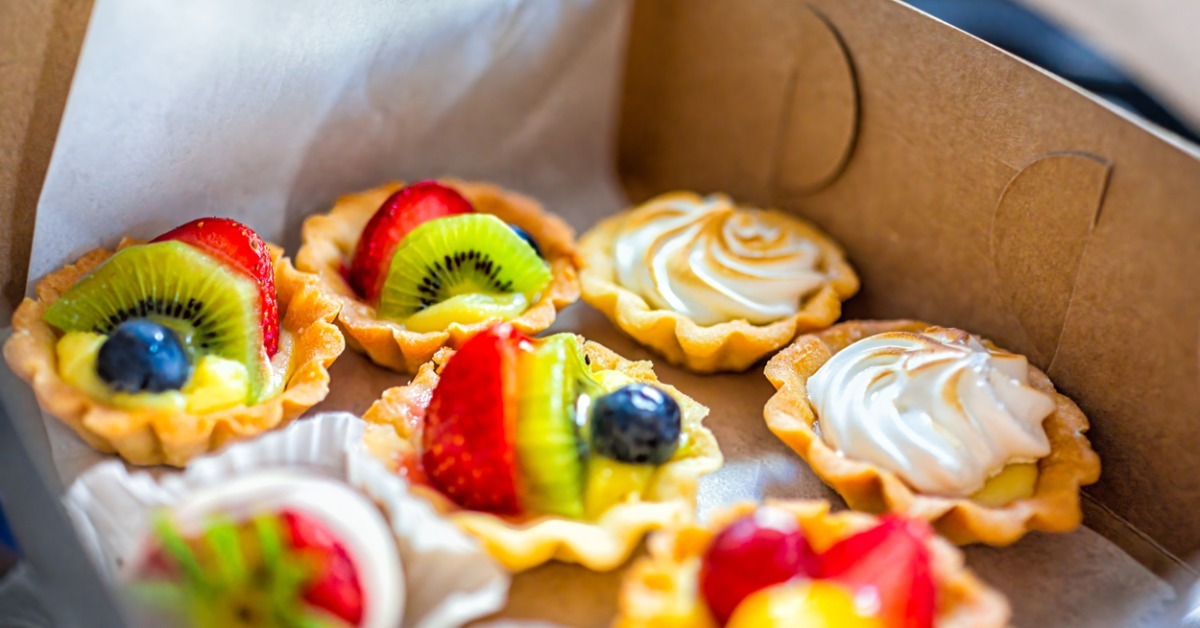 Assorted fruit tarts and cream tarts arranged in an open kraft bakery box lined with parchment paper for delivery. Assorted fruit tarts and cream tarts arranged in an open kraft bakery box lined with parchment paper for delivery.