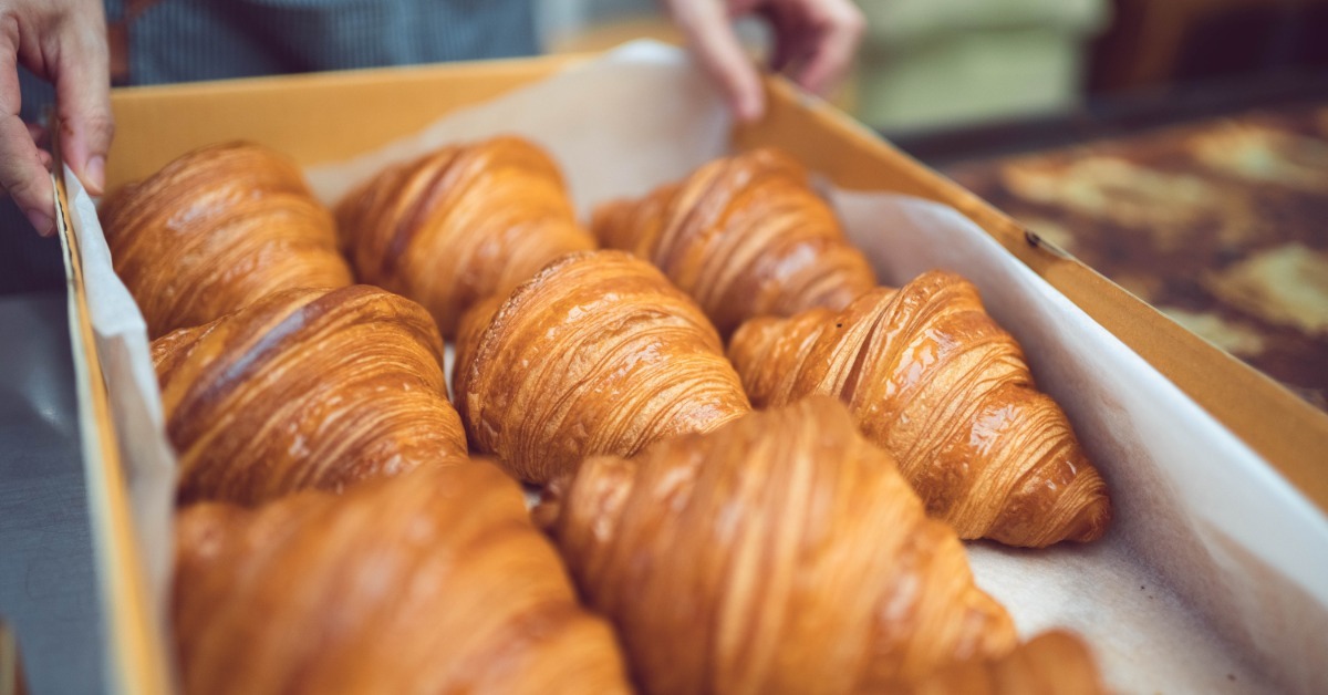 A baker holds a large kraft cardboard box filled with rows of fresh golden croissants lined with parchment paper. A baker holds a large kraft cardboard box filled with rows of fresh golden croissants lined with parchment paper.