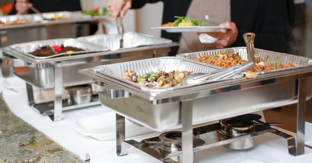 Guests serve themselves from foil-lined chafing dishes filled with catered food at a buffet table event. Guests serve themselves from foil-lined chafing dishes filled with catered food at a buffet table event.