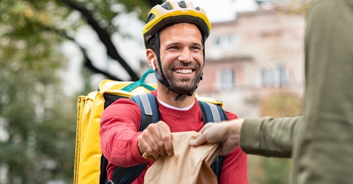 A smiling food delivery worker wearing a yellow helmet hands a brown paper bag to a customer outdoors.