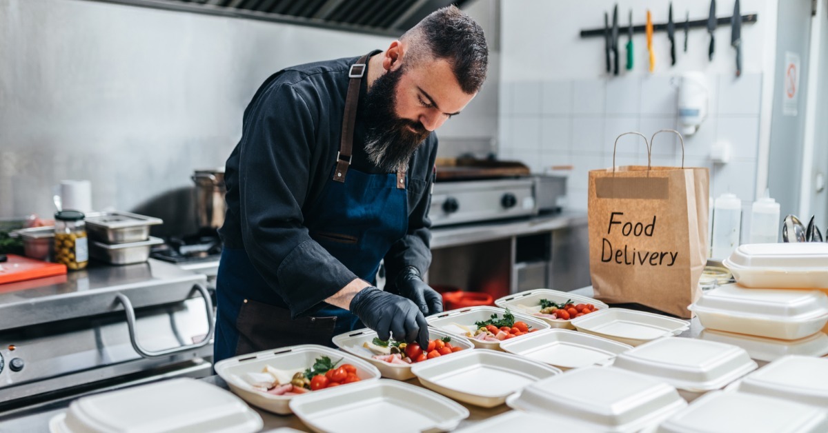 A bearded chef in a blue apron and gloves packs fresh salads into white takeout containers for food delivery.